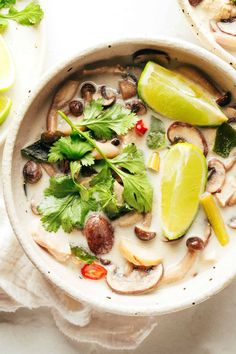 a white bowl filled with soup and garnished with cilantro, mushrooms, limes, and other vegetables