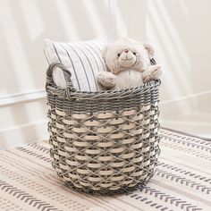 a teddy bear sitting in a basket on the floor next to a pillow and blanket