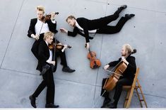 four men in suits are playing violin on the street while another man is sitting down