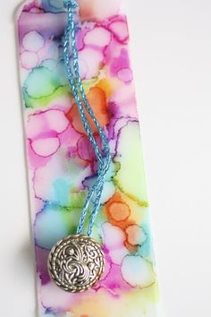 a colorful tie - dye bookmark with a silver medallion on it and a blue string