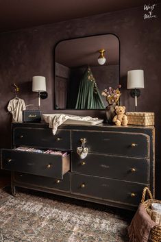 Dark purple nursery. Aged black vintage style dresser. Woman with brown hair wear a long black dress. There is a black mirror on the wall in between two black sconces with white lampshades. Nursery Ideas Storage, Preschooler Bedroom, Moody Nursery, West Of Main, Dark Nursery, Luxury Furniture Stores, Waiting For Baby, Changing Table Dresser, Baby Changing Table