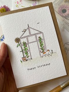 a hand holding up a card with a house and flowers on it that says happy birthday