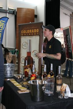 a man standing in front of a table with liquor bottles on it and people looking at them