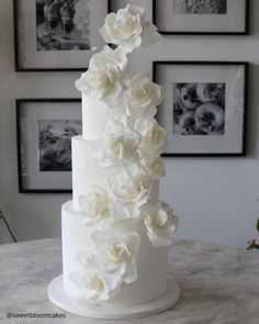 a three tiered white wedding cake with flowers on the top and bottom, sitting on a table in front of framed pictures