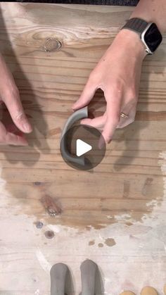 two hands holding something on top of a wooden table next to another person's feet