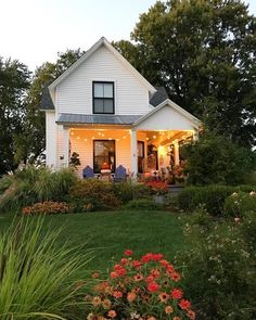 a white house with lots of flowers in the front yard and lights on it's porch