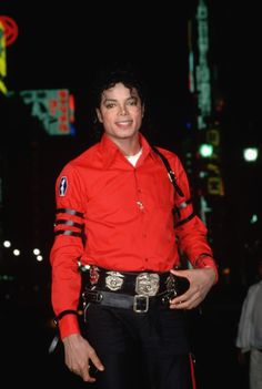 a man in red shirt and black pants standing on street at night with buildings behind him