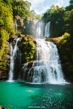 a man swimming in the water near a waterfall with green trees and rocks on either side