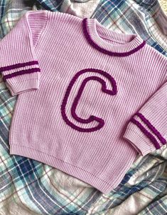 a pink sweater with the letter c on it sitting on top of a plaid blanket