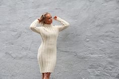 a woman standing in front of a gray wall wearing a white sweater dress and heels