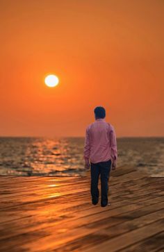 a man walking on a wooden pier towards the ocean with the sun setting in the background