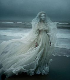 a woman in white dress walking on beach next to the ocean with her veil flowing