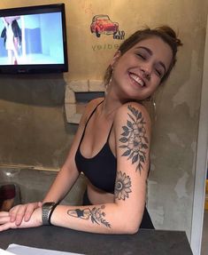 a woman sitting at a counter with tattoos on her arm and chest, smiling for the camera