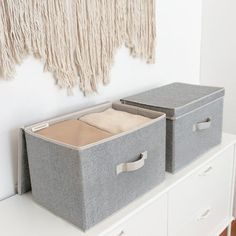 two storage bins on top of a white dresser next to a wall hanging with tassels