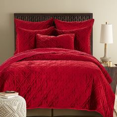 a bed with red comforter and pillows in a room next to a lamp on a table