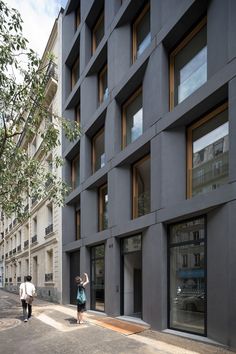 two people are walking down the sidewalk in front of a building with many windows on each side