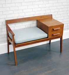 a small wooden desk with a green cushion on the top and bottom drawer, against a white brick wall