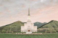 a large white church with a steeple in the middle of a green field and mountains behind it