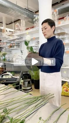 a woman holding a rose in her hand while standing next to shelves with flowers and plants