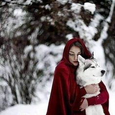 a woman in a red cloak holding a white husky dog on her lap while standing in the snow