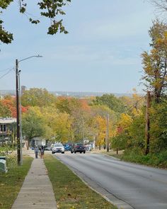 people are walking down the sidewalk in front of some parked cars and trees with fall colors