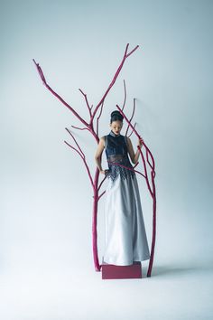 a woman in a dress standing next to a tree with red branches on the trunk