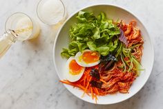 a white bowl filled with noodles and vegetables next to a glass of beer on a marble table