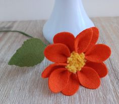 an orange flower sitting on top of a table next to a white vase with green leaves