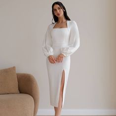 a woman in a white dress standing next to a couch and looking at the camera