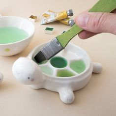 a hand is holding a paintbrush over some green liquid in a white bowl with two small bowls on the table