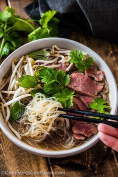 someone holding chopsticks over a bowl of noodles and meat with greens on top