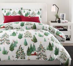 a bed with christmas trees on it and red pillows in the corner, next to a night stand