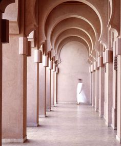 a person walking down a long hallway with arches and columns on both sides, in front of a pink wall