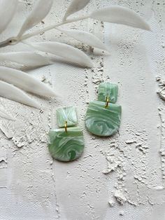 two green earrings with gold posts sitting on top of a white surface next to leaves