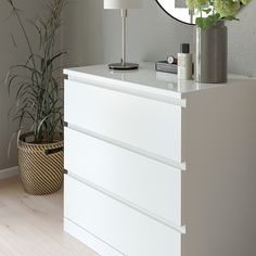 a white dresser sitting next to a mirror on top of a wooden floor near a potted plant