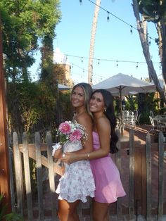 two women standing next to each other in front of a wooden fence with palm trees