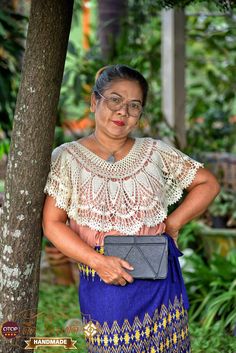 an older woman is standing next to a tree and holding a gray purse in her hands