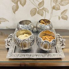 three silver bowls filled with different types of food on a metal tray next to a floral wallpaper