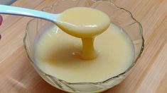 a spoon in a glass bowl filled with liquid on top of a wooden table next to a person's hand