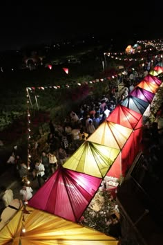 many colorful umbrellas are lit up in the night sky at an outdoor event with people standing around