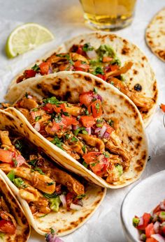 three tacos with chicken, tomatoes and cilantro on them next to a glass of beer