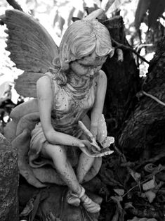 a black and white photo of a statue of a fairy sitting on the ground next to a tree