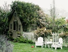 two lawn chairs sitting in front of a small wooden house with a white picket fence