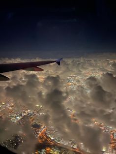 an airplane wing flying over the city lights in the night sky above some fluffy clouds