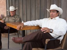 two men in cowboy hats sit at a table with their legs crossed and one man is sitting down