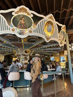 a woman standing in front of a carousel with paintings on the ceiling and people sitting at tables