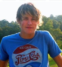 a young man in a pepsi cola t - shirt posing for the camera with trees in the background