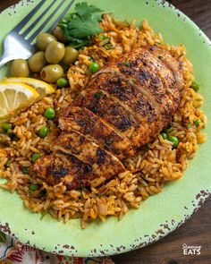 a green plate topped with chicken and rice next to olives, lemon wedges and a fork