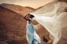 a woman in a blue and white dress standing on top of a rocky hill with her veil blowing in the wind