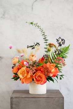 an arrangement of flowers in a white vase on a marble block with ferns and other foliage